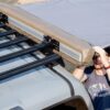 Person mounting a beige rooftop tent onto a car roof rack during a camping setup
