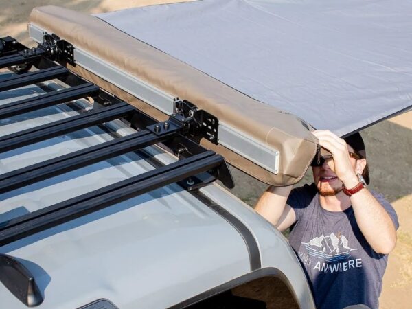 Person mounting a beige rooftop tent onto a car roof rack during a camping setup
