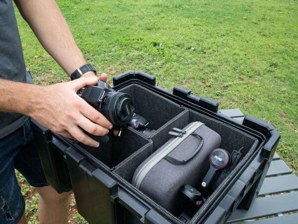 Person placing a DSLR camera into a padded, organized transport case outdoors on grass in a field bag of gear visible inside the case's foam compartments