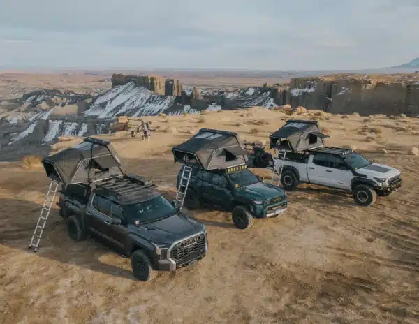 Three pickup trucks with rooftop tents parked on a sandy desert overlook, canyon walls in the distance.