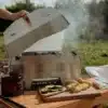 Open outdoor scene showing a Skotti portable grill on a wooden table with meat skewers and sliced vegetables cooking; a propane canister sits nearby and food prep boards are in view.