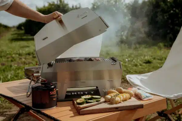 Open outdoor scene showing a Skotti portable grill on a wooden table with meat skewers and sliced vegetables cooking; a propane canister sits nearby and food prep boards are in view.