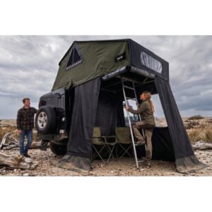 Two people set up a green rooftop tent on a black SUV in a rocky desert, with a ladder and camping chairs nearby.